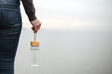 glass reusable water bottle hanging in a female hand on a background of cloudy sky