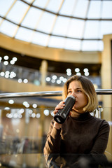 young European girl thoughtfully sits in a mall at sunset in and drinks from her hot thermos