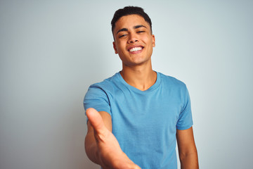 Obraz premium Young brazilian man wearing blue t-shirt standing over isolated white background smiling friendly offering handshake as greeting and welcoming. Successful business.