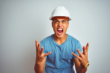 Young brazilian engineer man wearing security helmet standing over isolated white background crazy and mad shouting and yelling with aggressive expression and arms raised. Frustration concept.