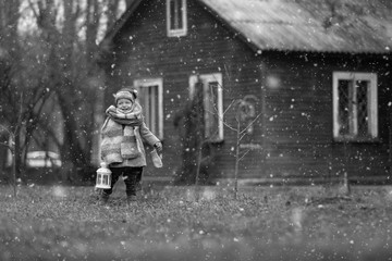 First snow. Little kid having enjoying countryside. Baby boy playing outdoors with vintage hand lamp, winter time old wooden house on background.