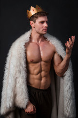 Portrait of a young bodybuilder with a crown on his head and a fur gown on his shoulders. Studio...