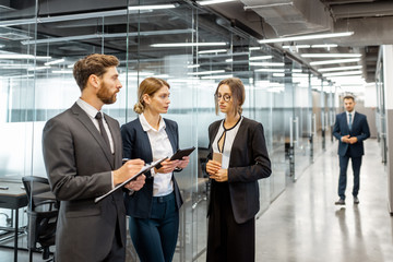 Group of business people strictly dressed in the suits meeting in the hallway of the modern office building, white-collar workers having informal discussion indoors