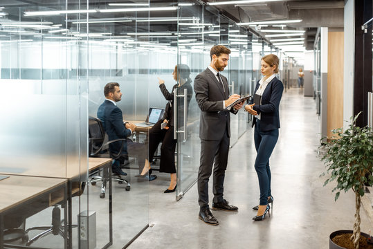 Business People Talking In The Hallway Of The Modern Office Building With Employees Working Behind Glass Partitions. Work In A Large Business Corporation