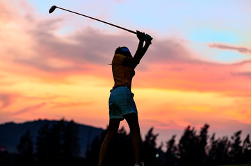 Silhouwttw of woman golf player in an action of backswing ready to hit a golf ball away to destination fairway at sunset in background