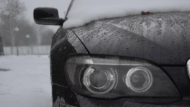 A headlamp closeup during snowfall in winter daytime. Bad weather conditions for traffic, blizzard. Danger for trips. Glassy black ice on the ground road. Snow covered automobile is in yard near house