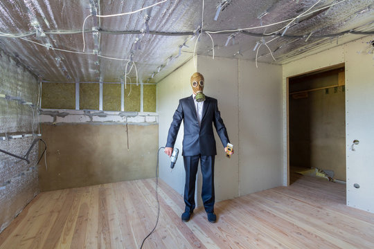 Strange man in businessman suit and gas protection mask inside a room under renovation works holding electric screwdriver and a level tools.