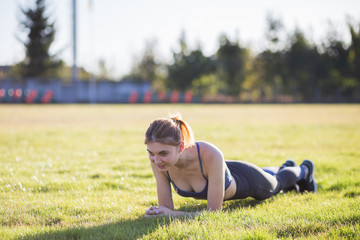 Young sportive woman in sports clothes training in field at sunrise. Girl standing in plank position on grass in a city park.