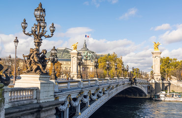 Famous Pont Alexandre III bridge on sunny autumn day. Behind is roof of Grand Palais with French flag waiving against clouds