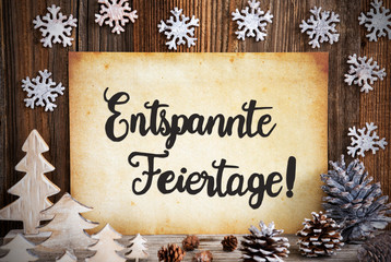 Fototapeta na wymiar Old Paper With German Text Entspannte Feiertage Means Merry Christmas. Christmas Decoration Like Tree, Fir Cone And Snowflakes. Brown Wooden Background