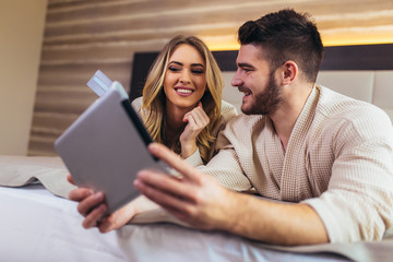 Happy couple using digital tablet while lying together in bed in hotel room.