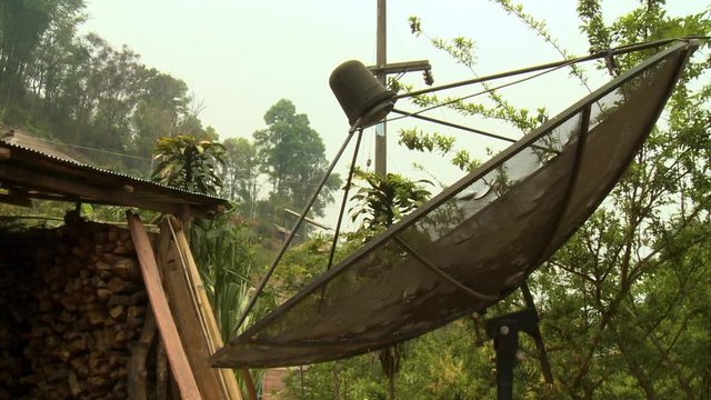 A steady closeup shot of a large satellite dish panel with a parabolic mesh plate erected near the entrance of a shed full of firewood.