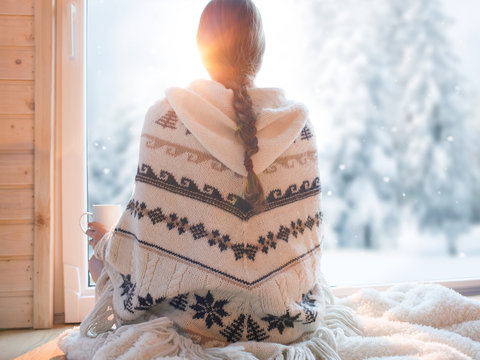 Woman wearing nordic style poncho sitting home by the window with cup of coffee, winter woods landscape outside