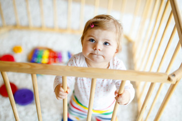Beautiful little baby girl standing inside playpen. Cute adorable child playing with colorful toys. Home or nursery, safety for kids. Alone baby waiting for mom