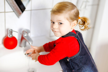 Cute little toddler girl washing hands with soap and water in bathroom. Adorable child learning cleaning body parts. Morning hygiene routine. Happy healthy kid at home or nursery.