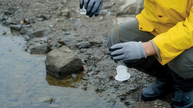 biotech expert in a respirator pours water from the river into a test tube for bacteriological analysis. Shakes to determine contamination. Ecology is modern.  Care about the world. close up
