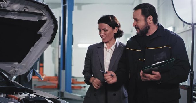 Charismatic mature mechanic in a uniform in a auto service center have meeting with the customer he showing the car and speaking about the problem