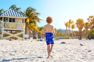 Blond little kid boy having fun on Miami beach, Key Biscayne. Happy healthy cute child playing with sand and running near ocean. Palms, security house and white sand