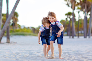 Two little kids boys having fun on tropical beach, happy best friends playing, friendship concept. Siblings brothers, twins fighting, running and jumping in family look with palms trees on background.