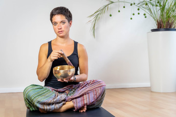 Relaxed woman with bronze singing bowl and clapper sitting on a yoga mat indoor
