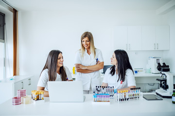 Three attractive smiling caucasian female lab assistants in sterile uniforms sitting and standing in lab while working on research. On lab table are test tubes, laptop, petri dishes and microscope.