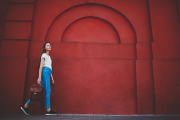 Fashionable young caucasian hipster girl walking at publicity area with trendy backpack in hand, attractive charming female student dressed in casual apparel going to university passing red wall