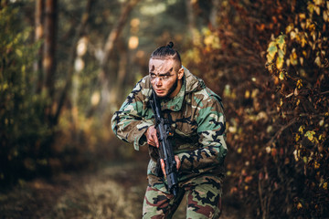 Fototapeta premium Portrait of a camouflage soldier with rifle and painted face playing airsoft outdoors in the forest