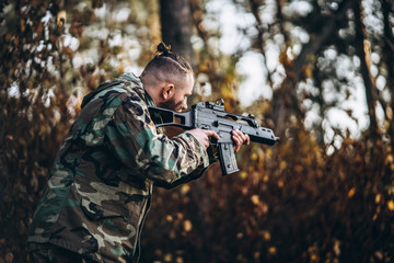 Portrait of a camouflage soldier with rifle and painted face playing airsoft outdoors in the forest. Side view