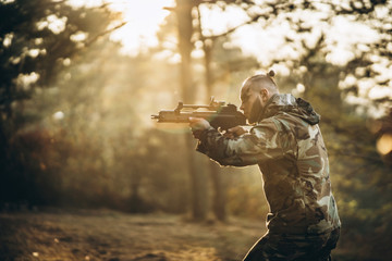 A camouflage soldier playing airsoft outdoors in the forest, sunset time, aiming at the rifle
