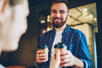 Half length portrait of cheerful young bearded man dressed in denim shirt smiling at camera while give tasty coffee to go to girlfriend during date in cafe.Positive hipster guy with hot beverage