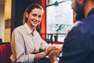 Half length portrait of happy hipster girl smiling at camera while holding hand of boyfriend on romantic date in cafe interior.Positive young woman spending leisure time with husband in coffee shop
