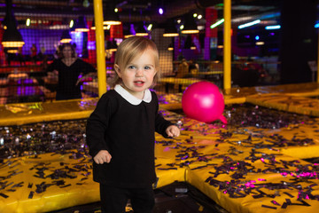 Adorable little girl looking at camera while standing near yellow mats with confetti and balloon during birthday party in family entertainment center trampoline park