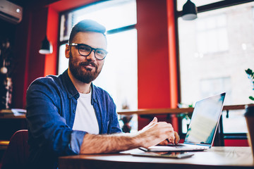 Portrait of bearded hipster guy in optical spectacles looking at camera during break from working remotely on modern netbook in cafeteria, handsome young man dressed in casual wear sitting with laptop