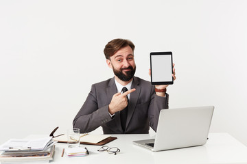 Indoor shot of young brunette man in formal clothes sitting at working table and looking to camera with ironic face, holding tablet pc and pointing on it with raised hand