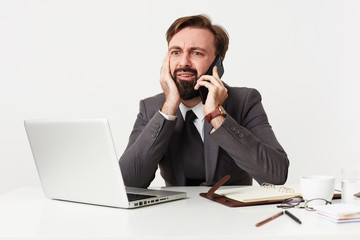 Bemused young brunette man with beard wearing formal clothes while working over white background, leaning head on raised hand and looking confusedly on screen of his laptop