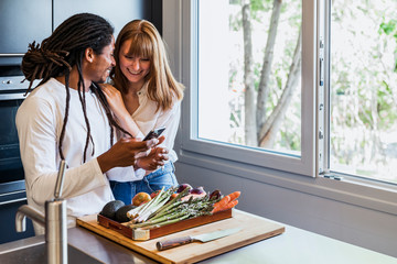 Girl leaning on the shoulder of a boy of different ethnicity in the kitchen looking at the mobile and smiling next to a window