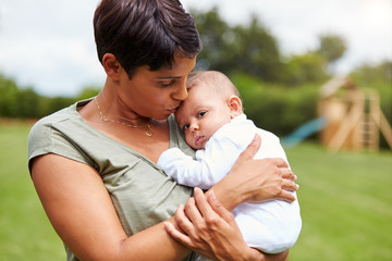 Loving Mother Holding And Kissing 3 Month Old Baby Daughter In Garden At Home