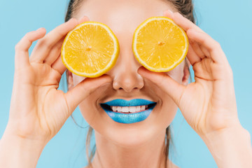 smiling beautiful woman with blue lips holding orange halves on eyes isolated on blue