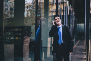 Old businessman in suit and and white beard having phone call while walking on street