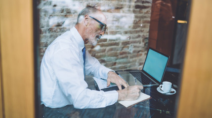 Senior male browsing laptop taking notes at cafe