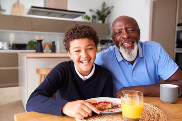 Portrait Of Smiling Grandfather Sitting In Kitchen With Grandson Eating Breakfast Before School