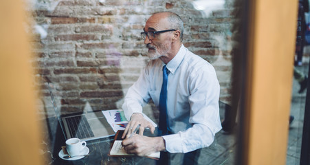 Senior man using laptop taking notes at cafe