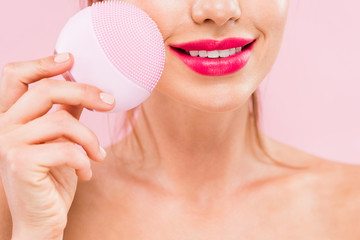 cropped view of naked beautiful woman with pink lips holding facial cleansing brush isolated on pink