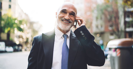 Cheerful senior businessman in formal outfit having phone call while walking on street