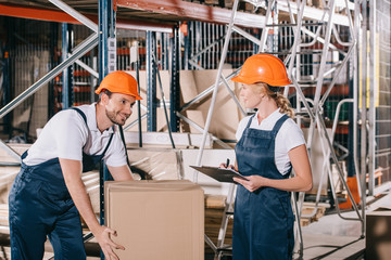 smiling loader holding cardboard box and looking at workwoman with clipboard