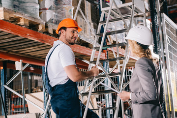 smiling warehouse worker standing on ladder and looking at businesswoman in helmet