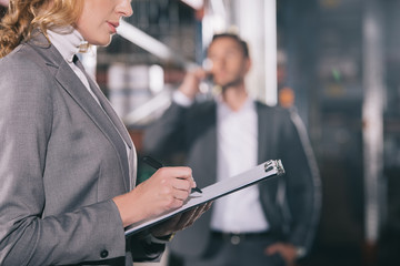 Obraz premium selective focus of businesswoman writing on clipboard near businessman talking on smartphone in warehouse