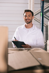 selective focus of smiling storekeeper holding clipboard while standing near cardboard boxes