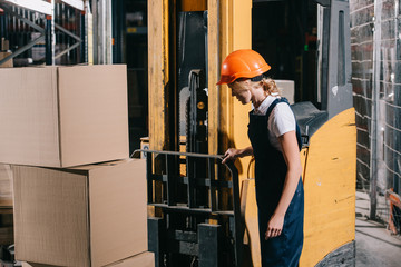 Workwoman in overalls and helmet standing near forklift loader in warehouse