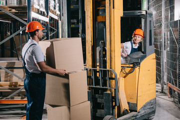 smiling workwoman sitting in forklift loader and looking at worker holding cardboard box